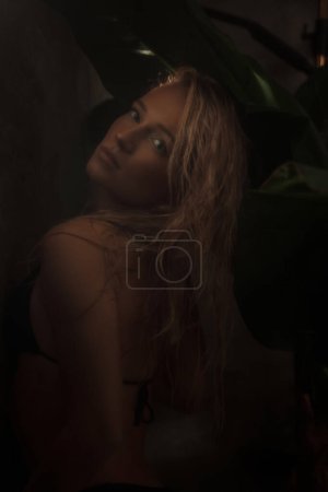Foto de Atractiva modelo femenina mostrando un concepto tropical mientras posa en un bikini negro en una ducha adornada con hojas de palma - Imagen libre de derechos