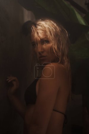 Photo for Attractive female model showcasing a tropical concept while posing in a black bikini in a shower adorned with palm leaves - Royalty Free Image