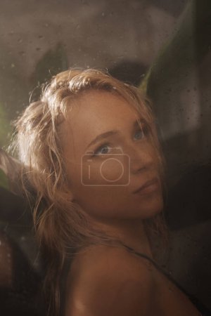 Photo for Close-up portrait of an enchanting blonde in a black swimsuit behind a shower glass surrounded by palm leaves, tropical concept, vacation concept in tropical countries - Royalty Free Image