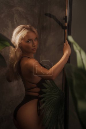 Foto de Modelo sexy en bikini negro posa en una ducha con una pared de textura gris, cubriéndose con una hoja de palma, concepto tropical, su rostro iluminado por la luz del sol - Imagen libre de derechos