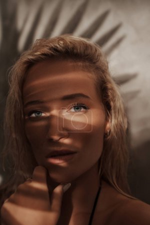 Photo for Close-up portrait of a stunning blonde in a black bikini, with palm branch shadows and sun-kissed skin, evoking a tropical vibe - Royalty Free Image