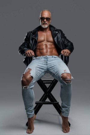 Photo for Handsome toned male model with a stylish gray beard with sunglasses, wearing a black leather jacket and ripped jeans, revealing his muscular physique and bare torso while posing on a studio chair - Royalty Free Image