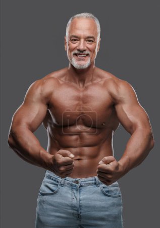 Téléchargez les photos : Bel homme plus âgé avec une barbe grise élégante, physique musculaire, jeans déchiré et torse nu, posant sur un fond gris - en image libre de droit
