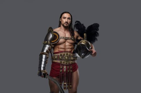 Photo for Powerful and attractive gladiator with a stylish beard and luscious locks wears ornate lightweight armor, clutching a gladius and feathered helmet as he stands confidently against a grey backdrop - Royalty Free Image
