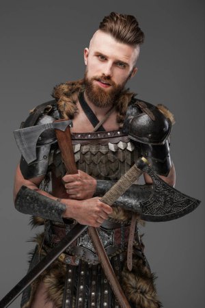 Foto de Un hombre vikingo imponente y rugoso con una barba tupida, revestido de piel de animal y armadura ligera, sosteniendo dos ejes sobre un fondo gris neutro - Imagen libre de derechos