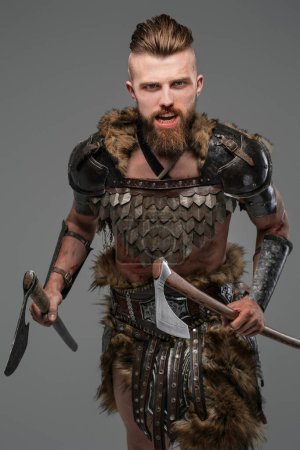 Photo for A fierce and bearded Viking warrior dressed in fur and light armor, wielding two axes against a gray background - Royalty Free Image