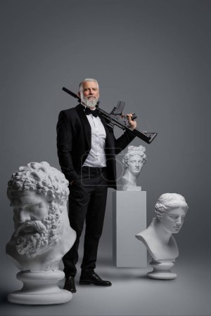 Photo for Suave and mysterious spy or secret agent with gray beard, dressed in a sharp suit confidently wields a gun, flanked by three classical sculptures, against a neutral gray background - Royalty Free Image