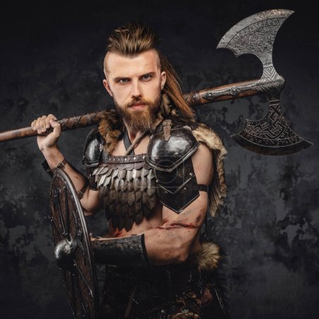 Téléchargez les photos : Un redoutable Viking barbu vêtu de fourrure et d'armure légère, brandissant une hache et un bouclier contre un mur gris texturé - en image libre de droit