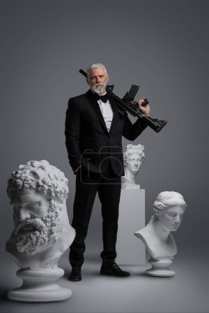 Téléchargez les photos : Élégant homme plus âgé se tient dans une pose confiante détendue sur un fond gris, tenant un pistolet, entouré de trois sculptures anciennes, vêtu d'un costume coûteux - en image libre de droit