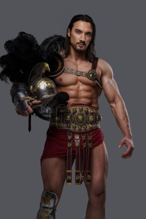 Téléchargez les photos : Gladiateur majestueux dans une armure légère élégante frappe une pose tout en tenant un casque sur un fond gris - en image libre de droit