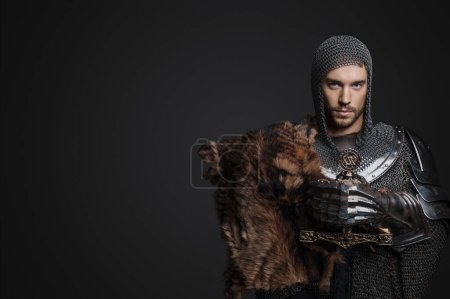 Photo for King wearing chainmail with reinforced steel plates, one shoulder covered by a fur pelt, strikes a regal pose while holding a sword against a neutral gray background in a portrait - Royalty Free Image
