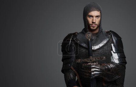 Téléchargez les photos : Portrait d'un beau roi en armure lourde, maille et couronne en acier, posant avec une épée sur un fond gris - en image libre de droit
