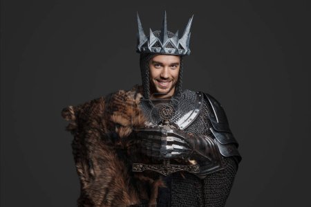 Foto de Retrato de estilo medieval de un joven rey guapo con armadura y cota de malla, sonriendo mientras sostiene una espada con una corona de acero y posa contra el gris - Imagen libre de derechos