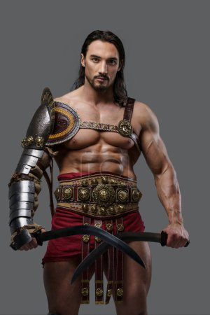 Foto de Elegante gladiador en elegante armadura ligera posa con dos gladios en la mano, sobre un fondo gris - Imagen libre de derechos