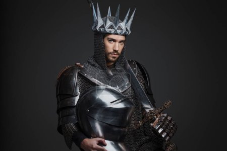 Téléchargez les photos : Portrait d'un beau roi en armure lourde, maille et couronne en acier, posant avec une épée sur un fond gris - en image libre de droit