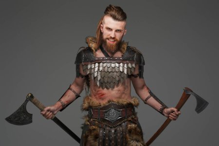 Photo for A fierce and bearded Viking warrior dressed in fur and light armor, wielding two axes against a gray background - Royalty Free Image