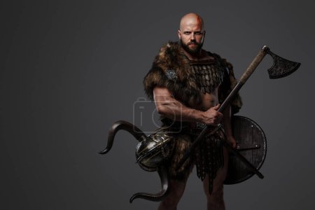 Téléchargez les photos : Un Viking robuste, chauve et barbu vêtu de fourrure et d'armure légère, avec un casque attaché à sa ceinture, tenant une hache et un bouclier sur un fond gris - en image libre de droit