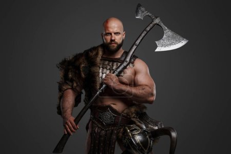 Téléchargez les photos : Un homme viking fort et intimidant à la barbe et à la tête chauve, vêtu de fourrure d'animal et d'armure légère, avec un casque suspendu à sa ceinture, brandissant une énorme hache sur un fond gris - en image libre de droit
