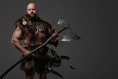 Foto de Un vikingo calvo y barbudo con una apariencia feroz que lleva piel y armadura ligera, llevando un hacha grande, contra un telón de fondo neutral - Imagen libre de derechos