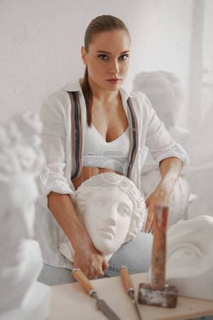 Téléchargez les photos : Image captivante d'un sculpteur habile, portant une chemise blanche et un jean avec des bretelles, dans une pose détendue avec un buste grec ancien - en image libre de droit