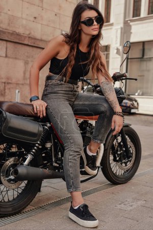 Foto de Elegante morena en jeans y una camiseta sin mangas, con gafas de sol y un tatuaje en el brazo, posa en su motocicleta retro en una vieja calle empedrada de Europa - Imagen libre de derechos