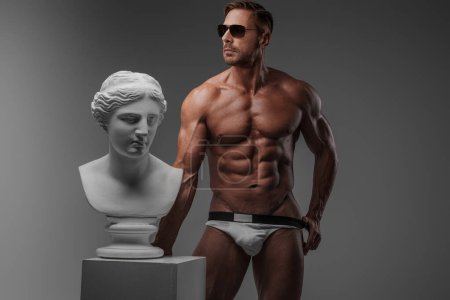 Foto de Atractivo modelo masculino muscular con gafas de sol y ropa interior se coloca con confianza junto al busto griego antiguo sobre un fondo gris - Imagen libre de derechos