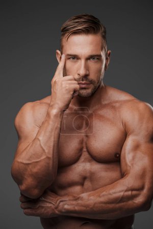 Foto de Un retrato de cerca de un modelo masculino musculoso posando pensativamente con un torso desnudo sobre un fondo gris - Imagen libre de derechos