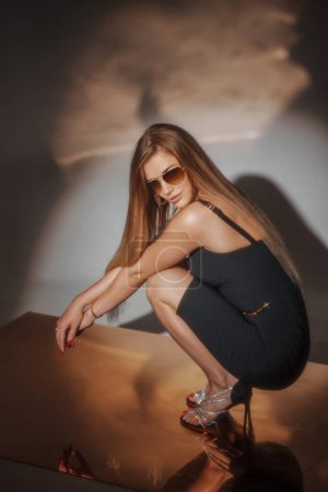 Téléchargez les photos : Superbe fille en robe de soirée noire et sunnies, frappant une pose assise sur la position accroupie sur un plancher d'or réfléchissant, avec un éclairage magnifique créant une toile de fond envoûtante - en image libre de droit