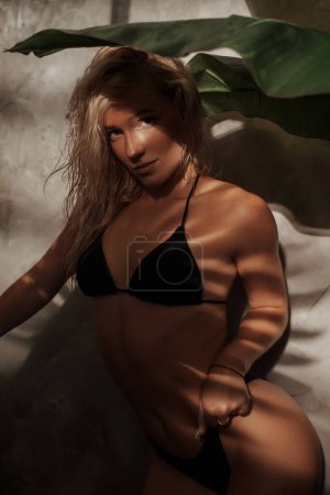 Téléchargez les photos : Blonde séduisante dans un bikini noir posant au milieu des feuilles de palmier, avec des ombres de branches de palmier et une peau ensoleillée, concept tropical - en image libre de droit