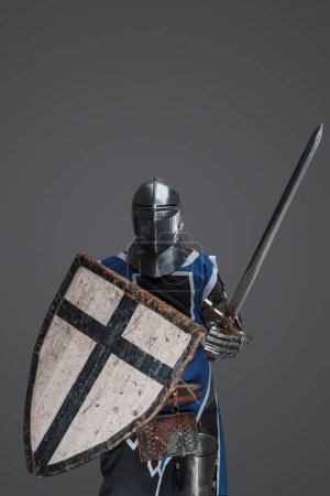 Foto de Brave medieval warrior dressed in armor and blue surcoat waving his sword energetically in battle, against a gray backdrop - Imagen libre de derechos