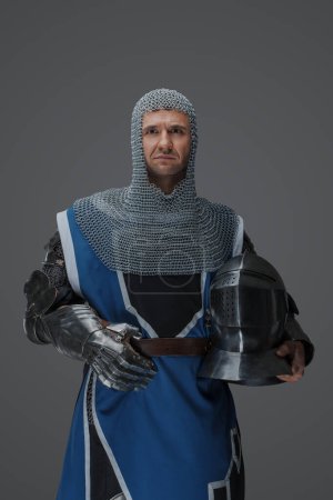 Téléchargez les photos : Garde royale vêtue d'armure médiévale et d'un survêtement bleu, avec coiffe à chaîne, tenant le casque à la main, sur un fond gris - en image libre de droit