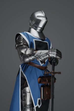 Téléchargez les photos : Un soldat médiéval vêtu d'un manteau bleu et d'une armure tenant une épée avec une pose immobile, semblable à une statue, sur un fond gris - en image libre de droit