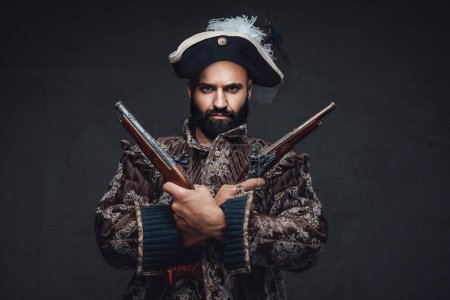 Photo for A pirate with a black beard holding two muskets, dressed in a vest and hat, against a dark textured wall - Royalty Free Image