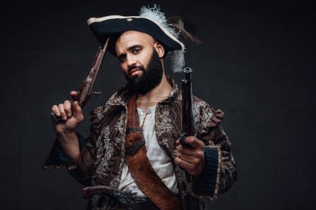 Téléchargez les photos : Pirate attrayant avec une barbe noire, vêtu d'un gilet et d'un chapeau, tenant deux mousquets contre un mur sombre texturé - en image libre de droit