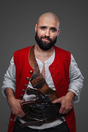 Foto de Pirata calvo de confianza con barba negra vestida con un chaleco rojo posando contra una pared gris - Imagen libre de derechos