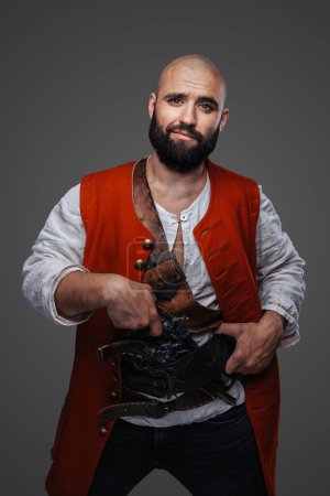 Téléchargez les photos : Pirate chauve confiant à la barbe noire vêtue d'un gilet rouge posant contre un mur gris - en image libre de droit