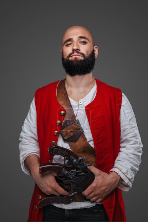 Photo for Confident bald pirate with black beard dressed in a red vest posing against a gray wall - Royalty Free Image