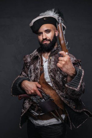 Photo for Pirate with a black beard holding two muskets, dressed in a vest and hat, against a dark textured wall - Royalty Free Image