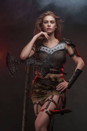 Téléchargez les photos : Une magnifique fille viking vêtue d'un haut de maille et d'une jupe en fourrure posant avec une hache à deux mains, éclairée par une lumière de jante rouge contre un mur gris texturé - en image libre de droit