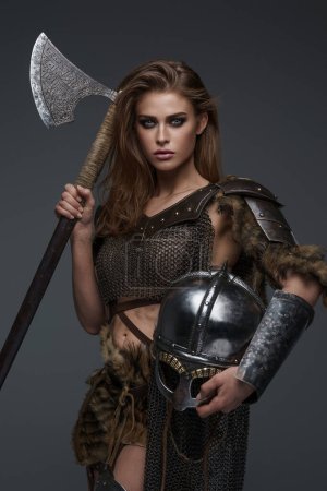 Impresionante modelo vikingo en armadura de cota de malla y hacha de piel y casco posando contra una pared gris