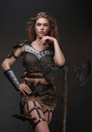 Impresionante mujer de inspiración vikinga posa con un hacha de dos manos contra una pared gris texturizada, con una parte superior de malla de malla y falda de piel