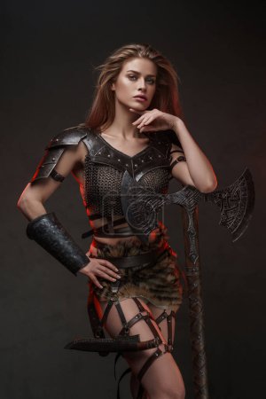 Foto de Impresionante chica vikinga vestida con una parte superior de malla de malla y falda de piel posa con un hacha de dos manos contra una pared gris texturizada - Imagen libre de derechos