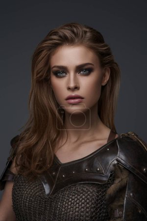 A close-up portrait of a model-like Viking woman wearing a chainmail top and fur posing against a grey background