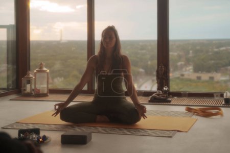 Téléchargez les photos : Femme en tenue de sport verte pratiquant le yoga et la méditation en pose de lotus sur fond de paysage urbain panoramique dans un appartement - en image libre de droit