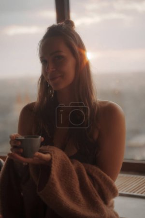 Téléchargez les photos : Fille profite d'une tasse de thé au milieu de l'atmosphère romantique d'un appartement de la ville, avec une toile de fond à couper le souffle de paysage urbain panoramique et un coucher de soleil incroyable - en image libre de droit