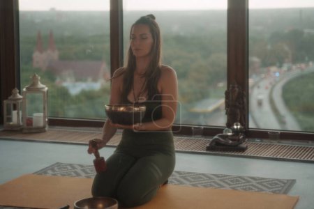Photo for A serene atmosphere as a woman engages in spiritual practices, meditating with a singing Tibetan bowl in a city apartment amidst a picturesque urban backdrop - Royalty Free Image