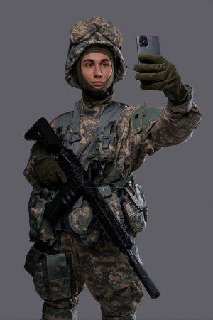 Foto de Joven en uniforme y casco sosteniendo un rifle y un smartphone, posando para una selfie o haciendo una videollamada sobre un fondo gris - Imagen libre de derechos