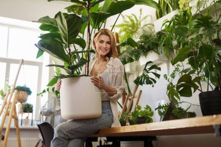 Foto de Chica sonriente en camisa blanca y jeans mostrando enorme planta en maceta en sala llena de vegetación - Imagen libre de derechos