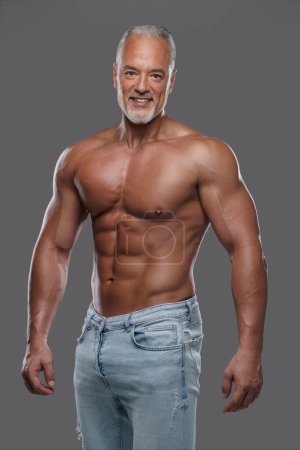 Téléchargez les photos : Homme plus âgé attrayant avec un physique musculaire, une barbe grise élégante et un jean déchiré, souriant et posant torse nu sur fond gris - en image libre de droit