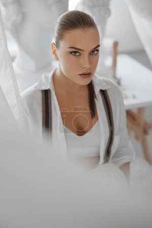 Téléchargez les photos : Portrait d'une sculptrice vêtue d'une chemise blanche déboutonnée et d'un jean à bretelles, tenant un buste grec ancien, posant assise sur une chaise dans son atelier de sculpteurs, entourée de sculptures - en image libre de droit